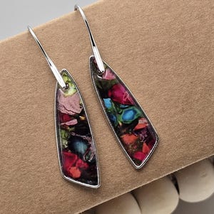 May include: A pair of silver dangle earrings with a colourful abstract design. The earrings are shaped like a teardrop and have a silver hook.