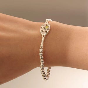 Puede incluir: Pulsera de tenis plateada con un dije de raqueta de tenis y una pequeña piedra amarilla. La pulsera está hecha de pequeñas cuentas redondas plateadas. Un accesorio elegante.