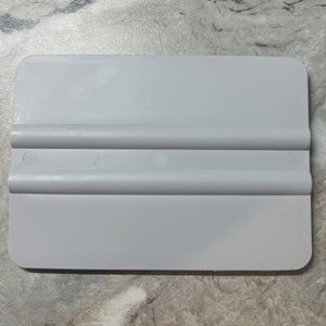 Puede incluir: Una escobilla de goma de plástico gris con una superficie lisa y dos bordes elevados. La escobilla tiene el número "2" impreso en ella.