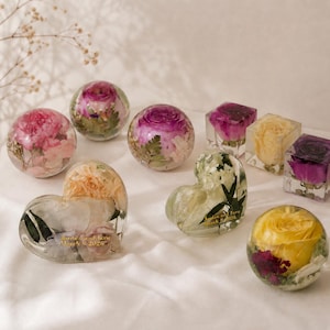 May include: A collection of clear resin keepsakes containing preserved flowers in spherical, heart, and cube shapes. Some pieces feature gold text reading Jordan and Sara March 8 2026. The items include pink, purple, yellow, and white floral arrangements.