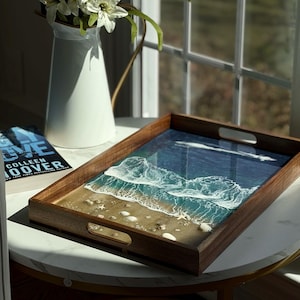 Puede incluir: Una bandeja de madera decorativa con una escena oceánica de resina. La bandeja tiene un marco y asas de madera oscura, con una representación detallada de olas, arena y conchas marinas. Un jarrón blanco con flores blancas está cerca.
