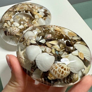 May include: Two clear resin candle holders filled with seashells and small stones. The candle holders are round and have a smooth, polished finish.
