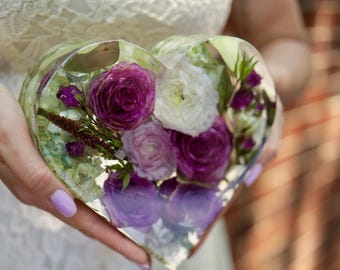 Puede incluir: Un pisapapeles de resina en forma de corazón lleno de un vibrante arreglo de flores moradas, blancas y lavanda. Las flores están encapsuladas en resina transparente, creando una pieza única y decorativa. El pisapapeles se sostiene frente a un vestido blanco.