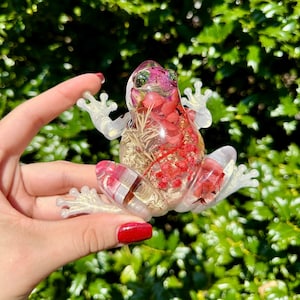 Könnte beinhalten: Eine durchsichtige Froschfigur aus Harz mit eingebetteten getrockneten Blumen und Blättern. Der Frosch ist hellrosa und hat einen weißen Umriss. Die Blumen sind rot und weiß.