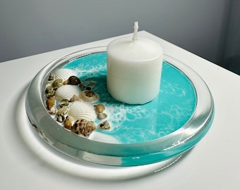 Puede incluir: Una vela blanca se encuentra en un plato de vidrio transparente con una base de resina azul turquesa. La base de resina tiene remolinos blancos y está decorada con pequeñas conchas.