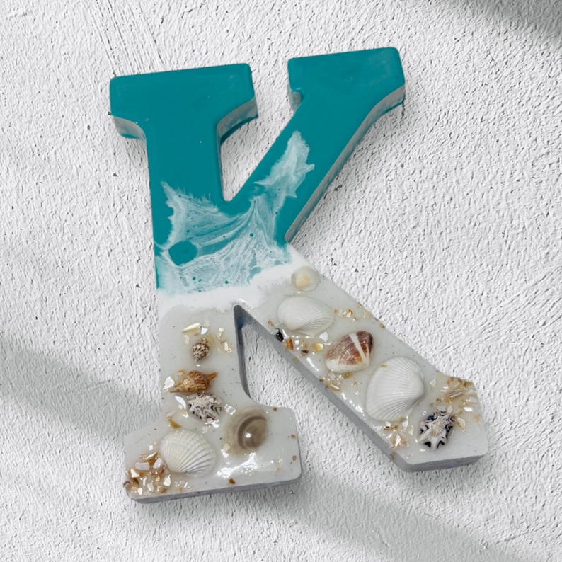 May include: A decorative letter "K" in teal and white, adorned with seashells and gold flakes. The top portion is teal, transitioning to white with a wave-like design. The lower section is white with embedded shells and gold accents.