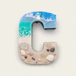 May include: A resin letter "C" with a beach theme. The top of the letter features blue and white resin, transitioning to turquoise and white. The bottom half is filled with sand, seashells, and starfish.