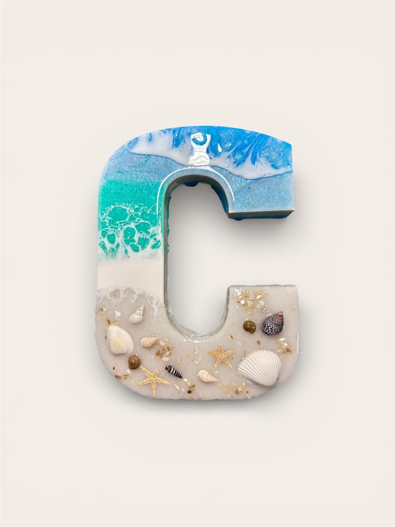 May include: A resin letter "C" with a beach theme. The top of the letter features blue and white resin, transitioning to turquoise and white. The bottom half is filled with sand, seashells, and starfish.