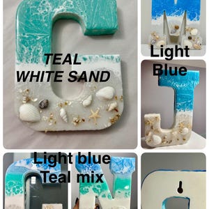 May include: Resin letters in various shades of blue and white, resembling ocean waves and sand. Each letter is adorned with seashells and starfish. The letters include the letters G, M, J, N, and R. The text "TEAL WHITE SAND" is visible.