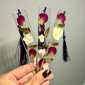 May include: Three clear resin bookmarks with dried flowers and purple tassels. Each bookmark features a different arrangement of dried rose petals, white flowers, and green leaves. The bookmarks are held together with a purple cord.