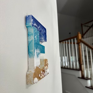 May include: A resin letter "E" mounted on a white wall. The top of the letter is blue, transitioning to white and turquoise, resembling ocean waves. The bottom section has a sandy texture with seashells.