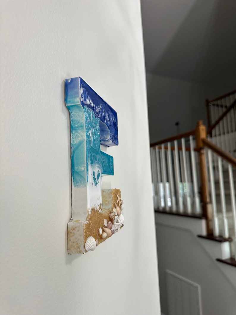 May include: A resin letter "E" mounted on a white wall. The top of the letter is blue, transitioning to white and turquoise, resembling ocean waves. The bottom section has a sandy texture with seashells.