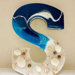 May include: A decorative letter "S" made of resin, featuring a beach theme. The top half is a gradient of blue and white, resembling ocean waves. The bottom half is white, embedded with seashells and starfish.