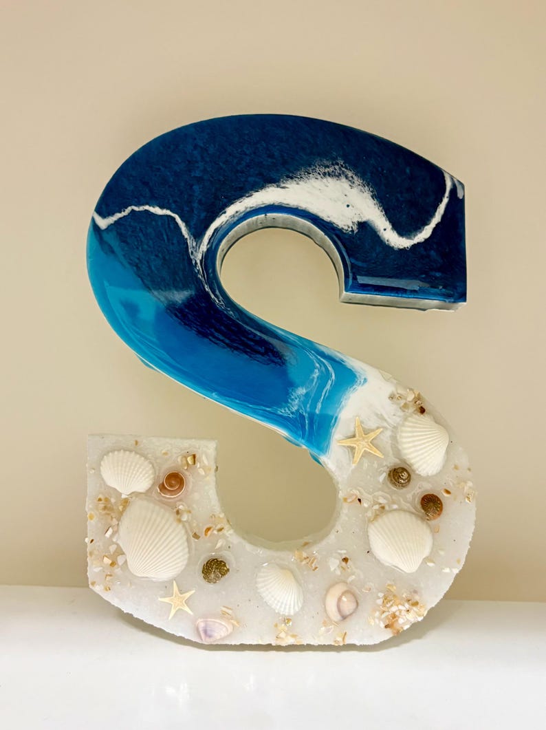May include: A decorative letter "S" made of resin, featuring a beach theme. The top half is a gradient of blue and white, resembling ocean waves. The bottom half is white, embedded with seashells and starfish.
