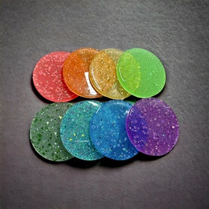 Puede incluir: Un juego de ocho posavasos redondos de resina llenos de purpurina. Los posavasos están dispuestos en círculo con los colores que pasan del rojo al verde al morado.
