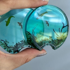 Puede incluir: Dos posavasos de resina azul turquesa con un diseño de peces y algas. Los posavasos tienen forma de semicírculo y tienen una pequeña muesca cortada en el lateral.