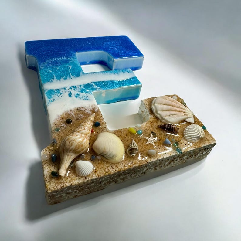 May include: A decorative letter "F" with a beach theme. The top is blue, resembling the ocean, transitioning to a sandy base adorned with seashells, sand, and small stones. The letter is approximately 6 inches tall.