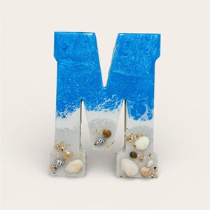 May include: A decorative letter "M" made of resin, featuring a blue and white ocean wave design at the top. The bottom portion is white with embedded seashells and sand, creating a beach-themed aesthetic.