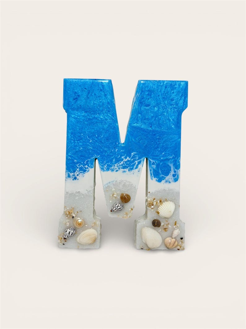 May include: A decorative letter "M" made of resin, featuring a blue and white ocean wave design at the top. The bottom portion is white with embedded seashells and sand, creating a beach-themed aesthetic.