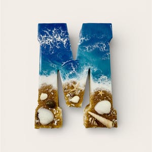 May include: A decorative letter "M" with a beach theme. The top of the letter is blue and white, resembling ocean waves. The bottom is tan, with seashells and sand. A unique coastal home decor item.