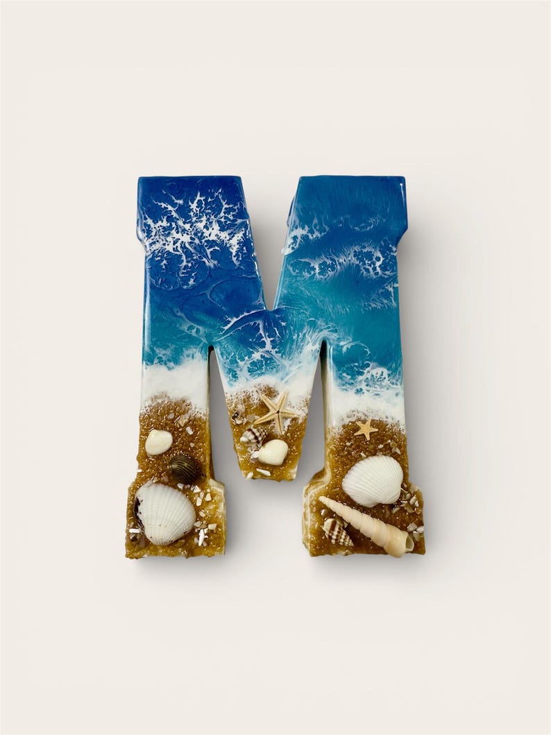May include: A decorative letter "M" with a beach theme. The top of the letter is blue and white, resembling ocean waves. The bottom is tan, with seashells and sand. A unique coastal home decor item.