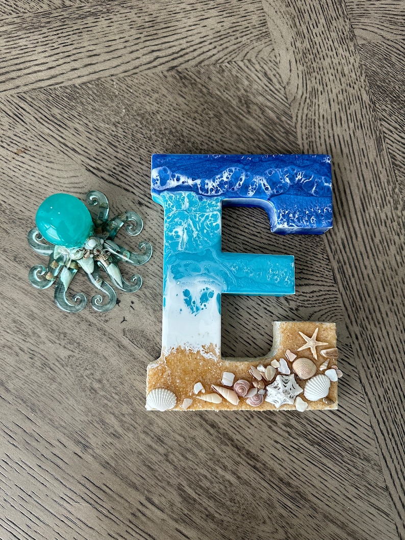 May include: A resin letter "E" with a beach theme, featuring shades of blue and white to mimic ocean waves. The base is adorned with sand, seashells, and a starfish. A glass octopus figurine with a turquoise orb is next to the letter.