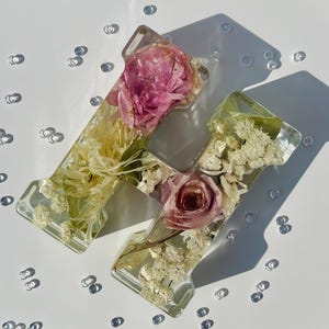 Könnte beinhalten: Ein dekorativer Buchstabe H aus klarem Kunstharz, eingebettet mit getrockneten rosa Rosen, weißem Schleierkraut und anderen cremefarbenen botanischen Elementen. Der Buchstabe ist von kleinen, verstreuten klaren Glasperlen umgeben.