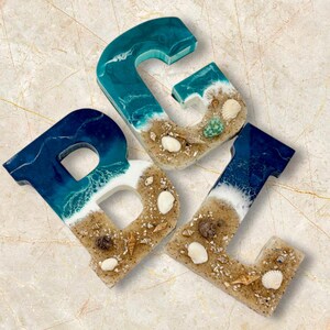 May include: Three resin letters, B, G, and L, are displayed. Each letter features a beach scene with blue and white waves, sand, and seashells. The letters are on a light-colored, marble-like surface.