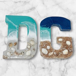 May include: Two resin letters, "D" and "G", with a beach theme. The "D" is turquoise and white, with seashells and sand. The "G" is blue and tan, also with seashells and sand. The letters are on a marble background.