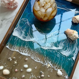 May include: A rectangular wooden serving tray featuring a resin art design that mimics a beach shoreline with sand, turquoise ocean water, and white foam waves. The tray is decorated with various small seashells and starfish embedded in the resin.