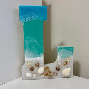 May include: A decorative letter "L" made of resin, featuring a beach-themed design. The top is blue, transitioning to turquoise, with a white "sand" base adorned with seashells and starfish. A unique coastal home decor item.