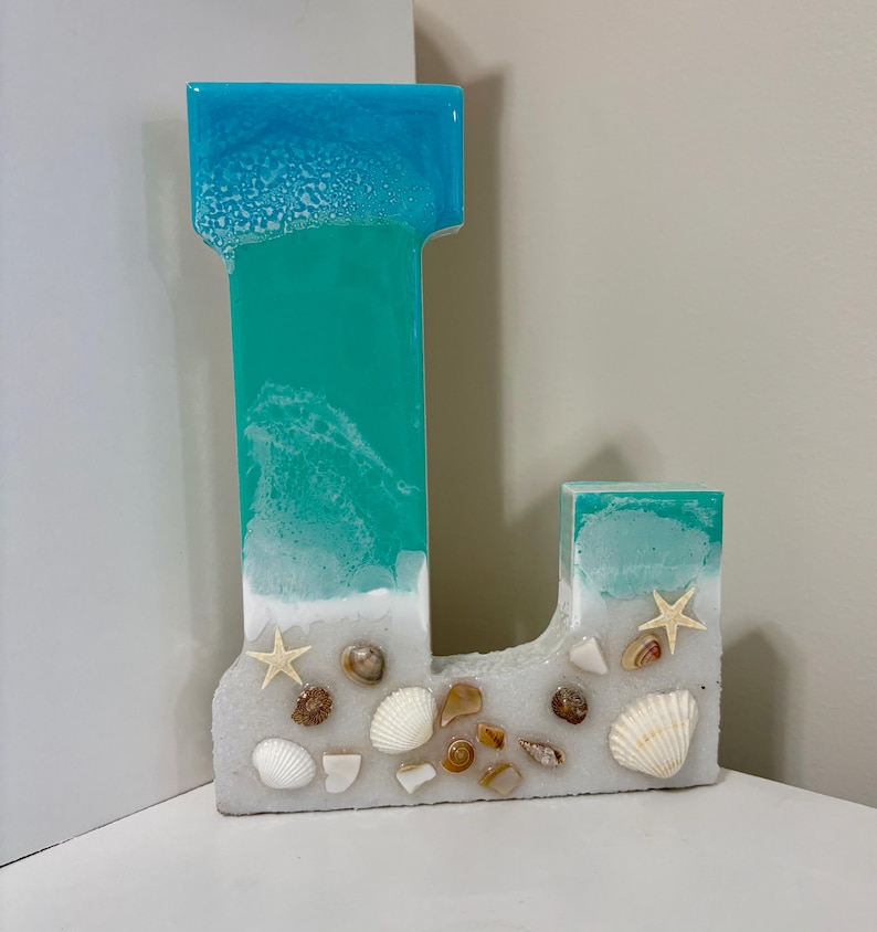 May include: A decorative letter "L" made of resin, featuring a beach-themed design. The top is blue, transitioning to turquoise, with a white "sand" base adorned with seashells and starfish. A unique coastal home decor item.