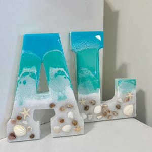 May include: Two resin letters, "A" and "J", with a beach theme. The letters are turquoise and white, resembling ocean waves, and are adorned with seashells and starfish. The letters are on a white surface.