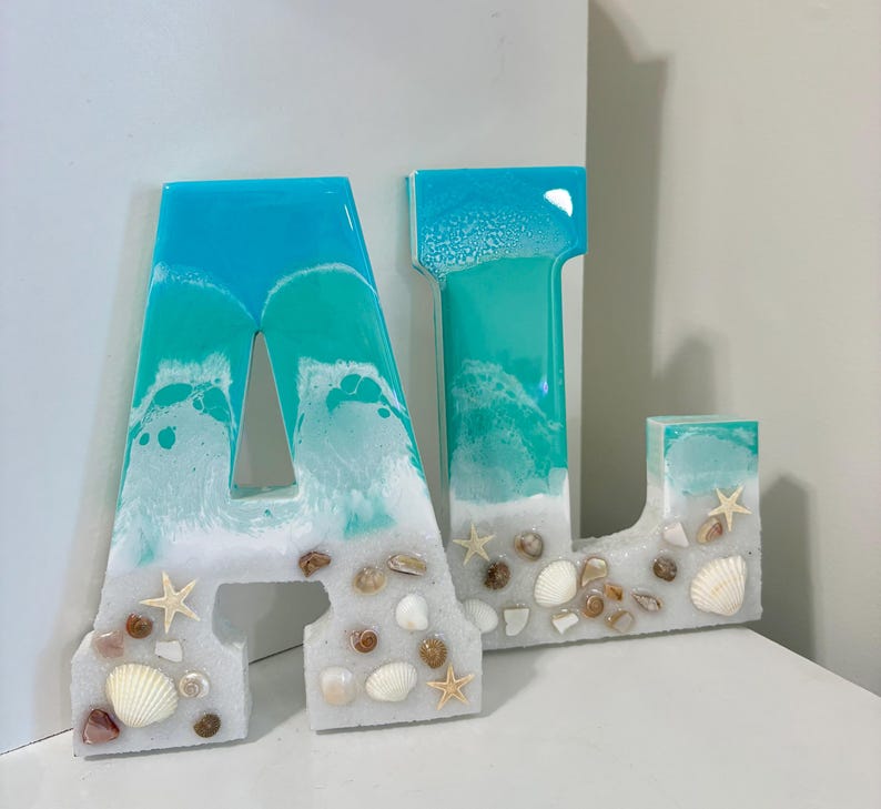May include: Two resin letters, "A" and "J", with a beach theme. The letters are turquoise and white, resembling ocean waves, and are adorned with seashells and starfish. The letters are on a white surface.