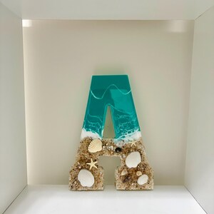 May include: A decorative letter "A" with a beach theme. The top portion is teal resin with white wave details. The bottom is sand-colored with seashells and starfish. A unique coastal home decor piece.