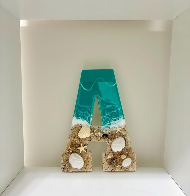 May include: A decorative letter "A" with a beach theme. The top portion is teal resin with white wave details. The bottom is sand-colored with seashells and starfish. A unique coastal home decor piece.