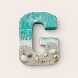 May include: A decorative letter "G" with a beach theme. The top is turquoise, resembling ocean waves, transitioning to white and sand-colored resin. Small seashells and a starfish are embedded in the sand-like base.