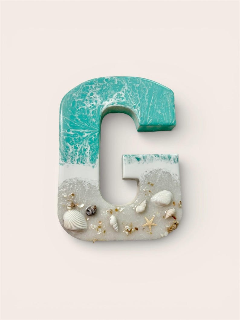 May include: A decorative letter "G" with a beach theme. The top is turquoise, resembling ocean waves, transitioning to white and sand-colored resin. Small seashells and a starfish are embedded in the sand-like base.