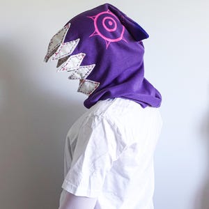 May include: A purple hooded cowl with a pink eye design and white diamond-shaped teeth. The cowl is made of soft fabric and is designed to be worn over the head. The hood is a unique accessory.