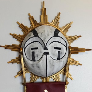 May include: A circular white mask with black painted details and a distressed finish, surrounded by a gold sunburst frame. A dark red fabric banner hangs from the bottom of the mask, featuring a gold painted triangular symbol in the center.