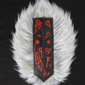 Könnte beinhalten: Ein schwarzes Holzschild mit rot bemalten Symbolen, vor einem Hintergrund aus weißem Kunstfell. Das Schild hat einen spitzen Boden und eine strukturierte Oberfläche. Die roten Symbole sind geometrisch und abstrakt.