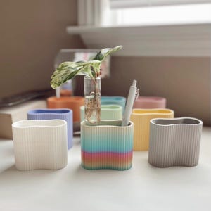 以下が含まれることがあります： 白、水色、薄い緑、オレンジ、グレーなど、さまざまな色の波打ったリブ付きのセラミックプランター6個セット。虹色のグラデーションデザインのプランターの1つに小さな植物が入っています。