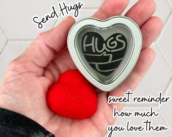 May include: A hand holding a small red felt heart and a silver heart-shaped tin with a black and white graphic that says "HUGS" inside. The text "Send Hugs" is written above the hand and the text "sweet reminder how much you love them" is written below the hand.