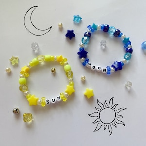 以下が含まれることがあります： 2つのビーズブレスレット。1つは星型のビーズと「SUN」の文字が入った黄色、もう1つは星型ビーズと「MOON」の文字が入った青色。散らばったビーズと太陽と月の絵も描かれています。