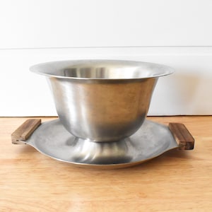 May include: A silver serving bowl with wooden handles sits on a matching silver plate. The bowl is round and has a wide, shallow base.