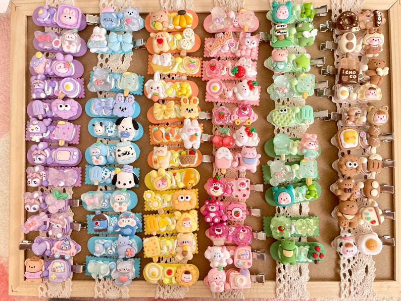 May include: A collection of 40 colorful hair clips featuring various cute cartoon characters and designs. The clips are arranged in rows on a white lace background. Each clip has a different design, including animals, food, and other whimsical themes.