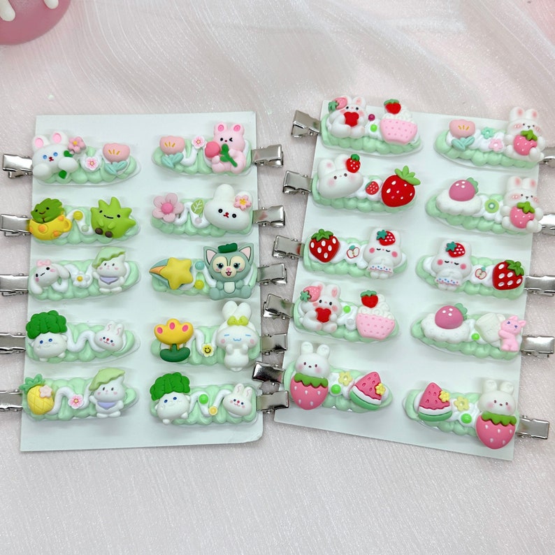 May include: A set of 24 colorful hair clips featuring various cute cartoon designs, including bunnies, strawberries, watermelons, flowers, and other whimsical characters. The clips are arranged on two white cards with green accents.