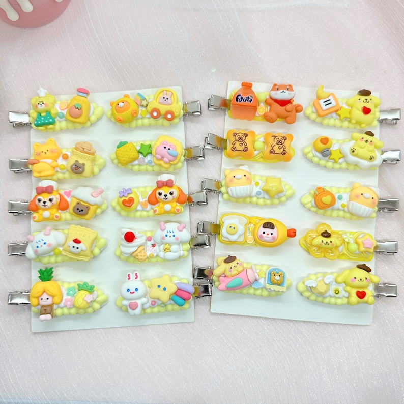 May include: A set of 20 yellow and white hair clips with various cartoon designs. The designs include a bear, a cat, a dog, a pineapple, a bunny, a star, a cloud, a car, a piece of bread, and a drink. The clips are on a white background.