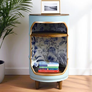 May include: A light blue barrel-shaped side table with a dark wood top and interior shelves. The interior features floral wallpaper. A framed black and white photo sits on top. The table is 24 inches tall.