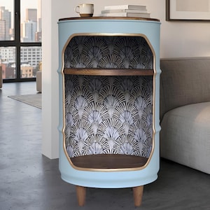 May include: A light blue cylindrical metal barrel cabinet with a dark wood top and two interior shelves. The interior features a black and white fan-shaped pattern, and the cutout opening is trimmed with a gold-colored border. It stands on three short wooden legs.